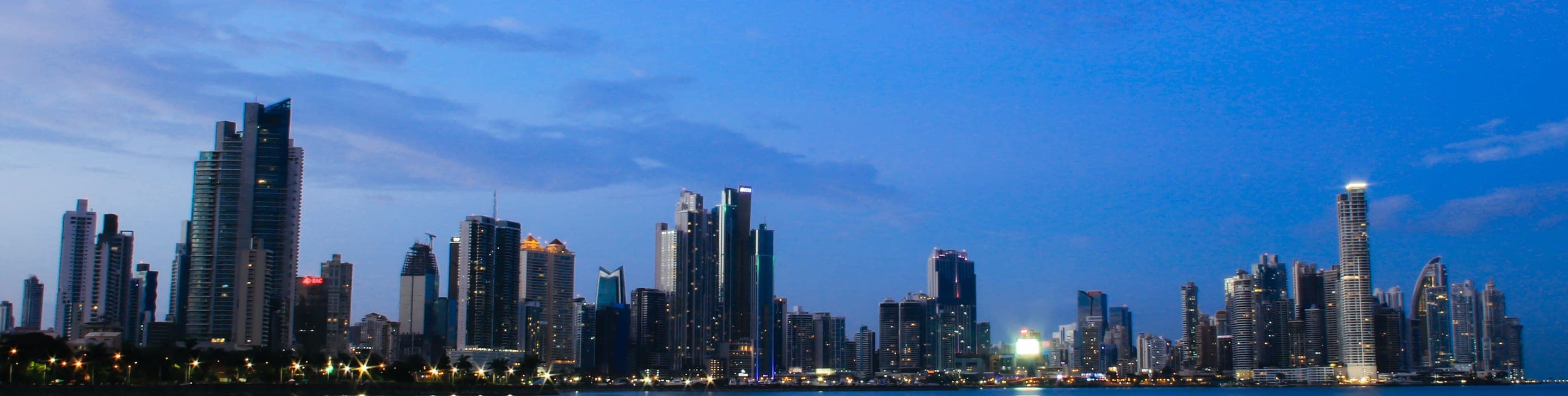 A city skyline of illuminated high-rise towers reflected in calm water at dusk in Panama